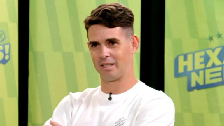 Oscar Emboaba com camiseta branca e microfone de lapela cruzando os braços durante entrevista no estúdio do Hexa Neles com fundo verde e logotipo ao lado