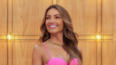 Patrícia Poeta sorri com vestido rosa e colar dourado diante de painel de madeira iluminado em estúdio de TV