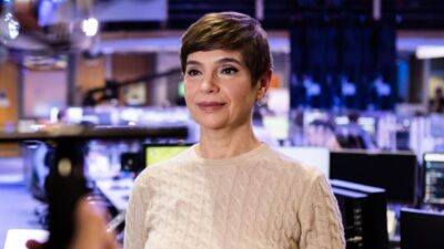 Renata Lo Prete com blusa clara em estúdio de jornalismo da Globo durante gravação na redação da emissora