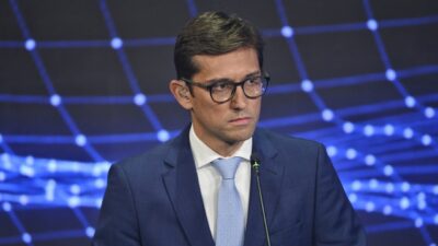 Rodolfo Schneider de terno azul e gravata clara durante debate em estúdio com painel digital azul ao fundo