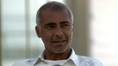 Romário com camisa branca estampada durante entrevista em ambiente iluminado com expressão séria e olhar lateral