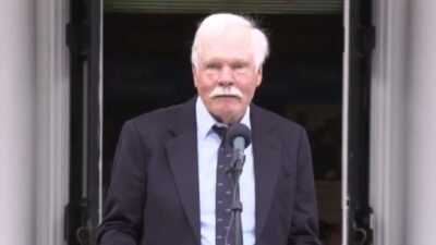 Ted Turner fala ao microfone diante de uma porta aberta usando terno escuro e gravata azul