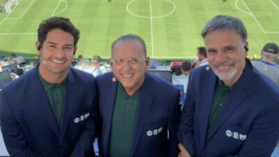 Alexandre Pato, Galvão Bueno e Mauro Beting sorrindo em cabine de transmissão com gramado de estádio ao fundo
