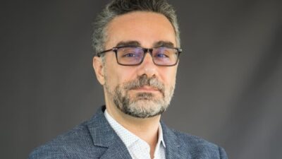André Marsiglia de óculos e blazer cinza olha para a câmera diante de fundo cinza com barba grisalha