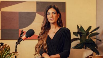 Andréia Sadi posa ao lado de microfone em estúdio de podcast com quadro geométrico e plantas ao fundo