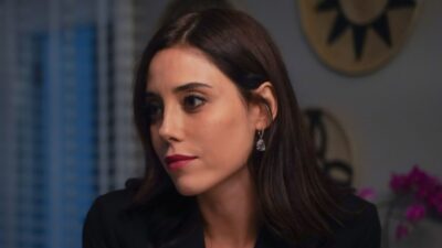 Asya Yilmaz (Cansu Dere) de blazer preto observa alguém com expressão séria em ambiente interno na novela Iludida