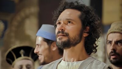Daniel (Gabriel Gracindo) com expressão apreensiva observa à frente cercado por homens em cena de A História de Daniel