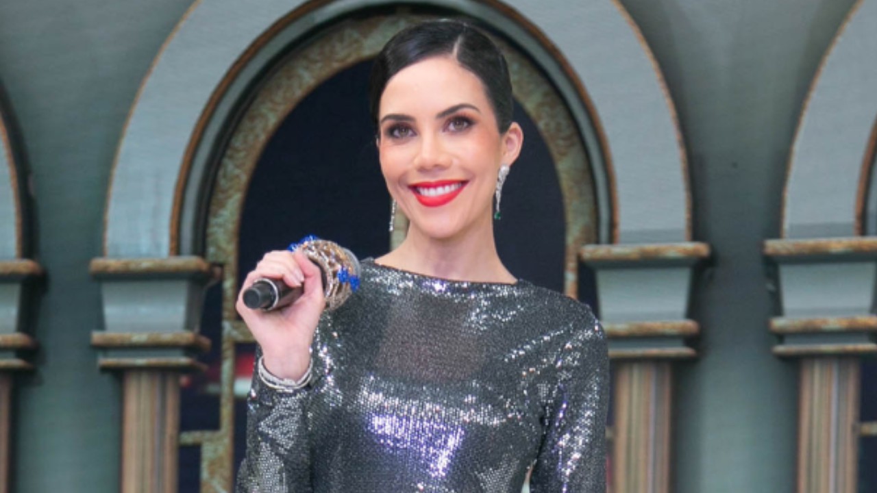 Daniela Albuquerque de vestido prateado brilhante sorri com microfone na mão no palco do Sensacional