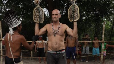 Ed Stafford segura dois artefatos indígenas durante ritual cercado por integrantes de comunidade na série Ed Stafford: Rituais Extremos