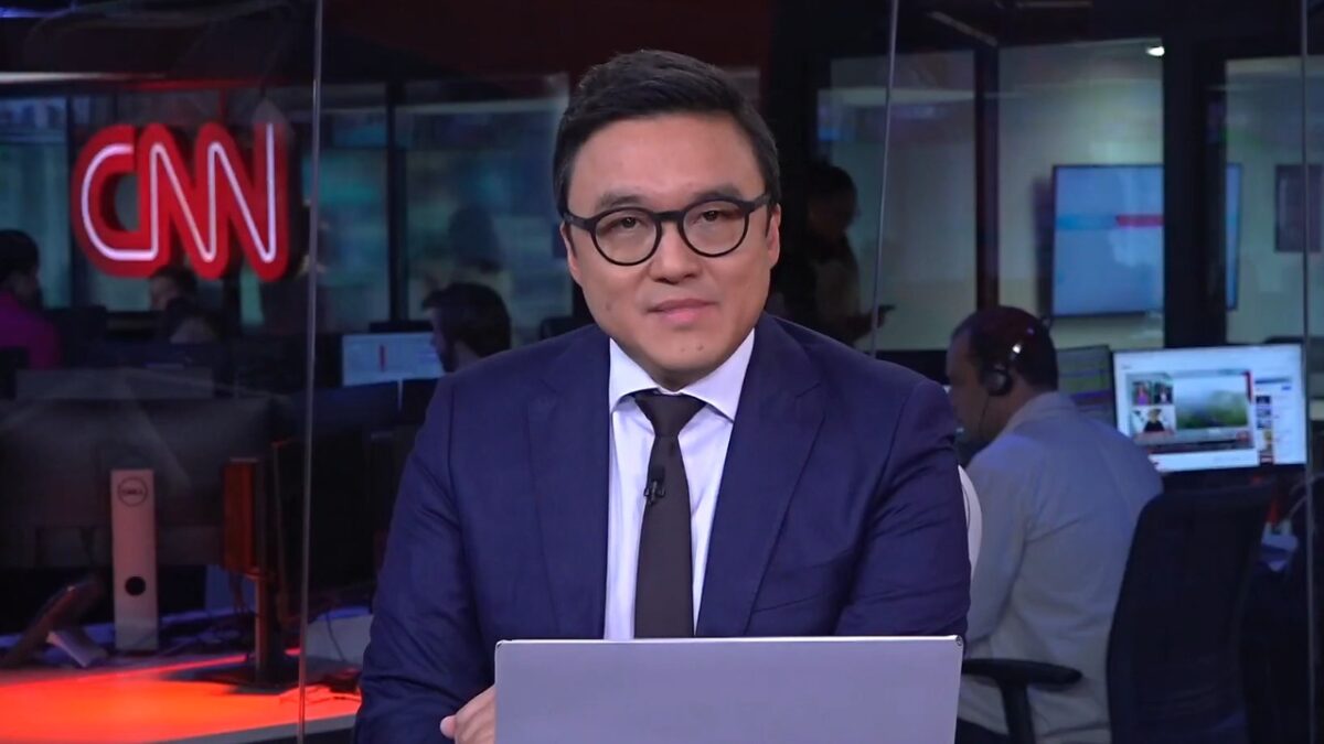 Fernando Nakagawa de terno azul e gravata escura diante de notebook na redação da CNN Brasil com monitores ao fundo
