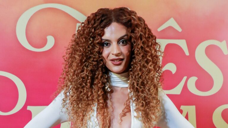 Gabriela Loran posa diante de painel promocional com cabelo cacheado volumoso e roupa branca em evento de lançamento