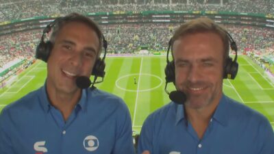 Gustavo Villani e Roger Flores de headset durante transmissão da Copa do Mundo com campo e arquibancadas ao fundo
