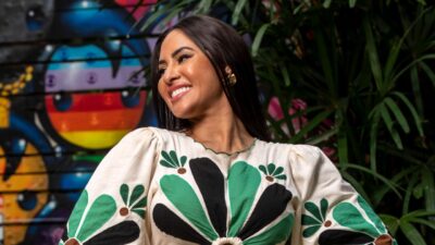 Isabelle Nogueira sorri com vestido estampado em verde e preto diante de mural colorido e folhagens em ambiente interno