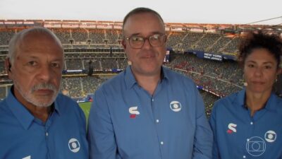 Junior, Everaldo Marques e Cristiane de camisas azuis em cabine de transmissão com estádio ao fundo na Copa do Mundo