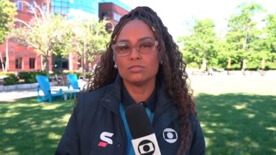 Karine Alves com microfone da TV Globo durante entrada ao vivo em área externa com árvores e edifícios ao fundo