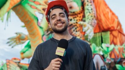 Lucas Selfie sorri com capacete vermelho e microfone na mão diante de carro alegórico colorido com dragão ao fundo