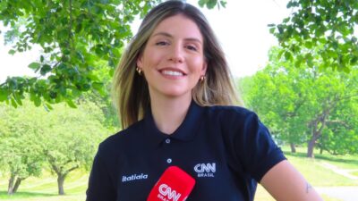 Ludmila Candal sorri segurando microfone da CNN Brasil em área arborizada com árvores e gramado ao fundo