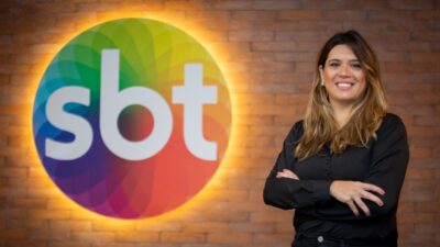 Marcela Marchi de blusa preta com braços cruzados diante de logotipo do SBT em parede de tijolos iluminada