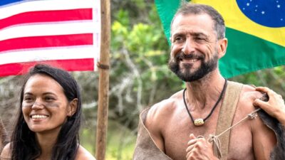Marina Fukushima e René Murad sorriem ao ar livre diante das bandeiras dos Estados Unidos e do Brasil em Largados e Pelados - Campeões do Mundo