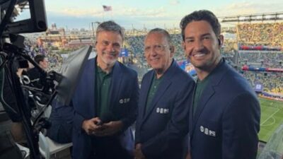 Mauro Beting, Galvão Bueno e Alexandre Pato sorriem em cabine de transmissão com estádio ao fundo durante jogo de futebol