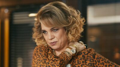Pilar (Isabel Teixeira) com expressão tensa e olhar voltado para baixo, segurando o próprio ombro, usando blusa de estampa animal print, relógio e pulseiras douradas, em ambiente interno da novela Quem Ama Cuida