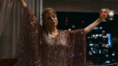 Pilar (Isabel Teixeira) ergue taça e abre os braços com vestido estampado diante de janela com vista noturna da cidade