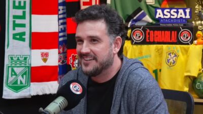 Raphael Rezende sorri diante de microfone em estúdio de podcast com camisas e bandeiras de futebol ao fundo