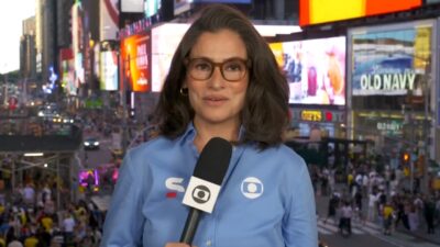 Renata Vasconcellos de camisa azul e óculos com microfone da Globo em frente aos painéis luminosos da Times Square em Nova York