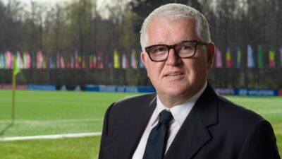 Romy Gai de terno escuro e gravata posa diante de campo de futebol com bandeiras ao fundo usando óculos de armação preta