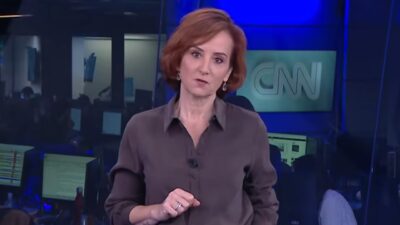 Thaís Herédia de blusa cinza durante apresentação em estúdio da CNN Brasil com redação ao fundo