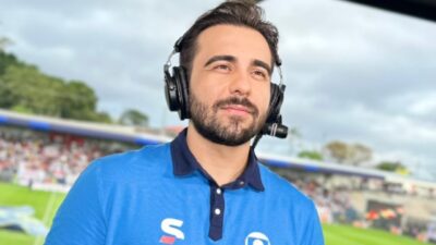 Vinícius Rodrigues de camisa azul e headset observa o campo de futebol durante transmissão esportiva da Globo