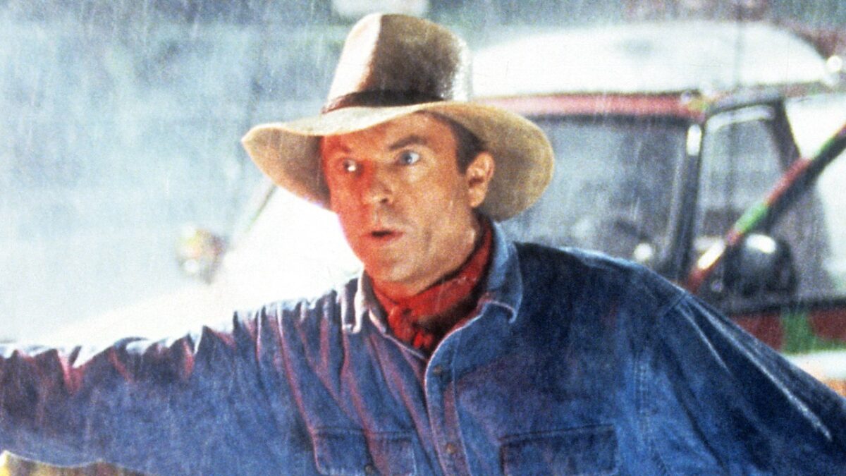 Alan Grant (Sam Neill) de chapéu, camisa jeans e lenço vermelho com expressão de alerta sob chuva em Jurassic Park