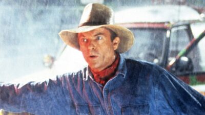 Alan Grant (Sam Neill) de chapéu, camisa jeans e lenço vermelho com expressão de alerta sob chuva em Jurassic Park