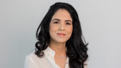 Ana Soares sorri usando camisa branca e colar discreto diante de fundo cinza claro em retrato de estúdio