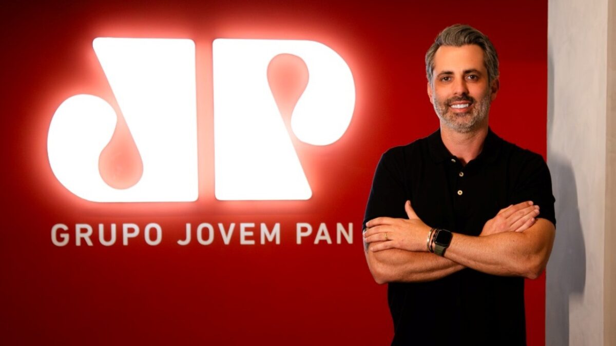 Arthur Ingnez de camisa polo preta com braços cruzados sorri diante do logotipo iluminado do Grupo Jovem Pan