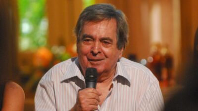 Benedito Ruy Barbosa sorri enquanto segura um microfone com a mão direita, veste camisa listrada clara e está em ambiente interno com fundo desfocado e iluminação quente