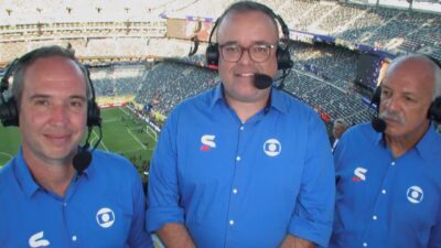 Caio Ribeiro, Everaldo Marques e Junior de camisas azuis com headsets em cabine de estádio com campo ao fundo