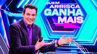 Celso Portiolli de blazer escuro segura microfone e aponta para o telão com logo do quadro Quem Arrisca Ganha Mais sorrindo no estúdio