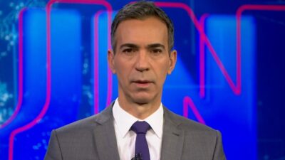 César Tralli de terno cinza e gravata azul olha para a câmera no estúdio do Jornal Nacional com painel azul ao fundo