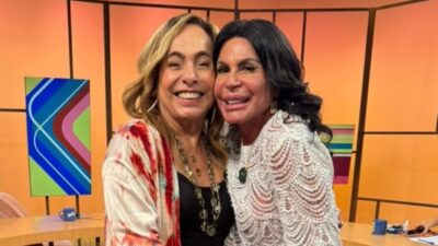 Cissa Guimarães e Gretchen sorriem abraçadas no estúdio do Sem Censura, Cissa veste blusa preta com kimono estampado e colares, Gretchen usa blusa branca rendada, ao fundo há mesas, canecas e painéis coloridos do cenário do programa