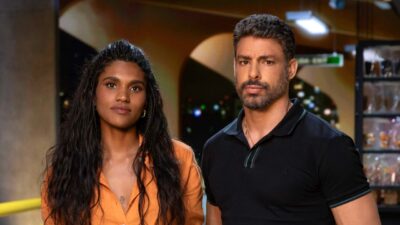Cris (Mariana Sena) de blusa laranja e Maurício (Cauã) de camisa polo preta lado a lado com expressão séria em ambiente interno iluminado da série Jogada de Risco
