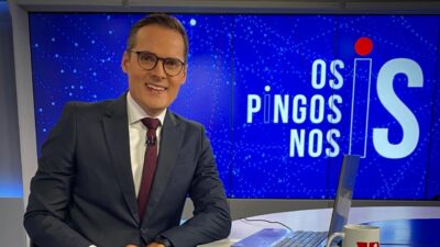 Daniel Caniato de terno e gravata sorri na bancada do Os Pingos nos Is com telão do programa ao fundo
