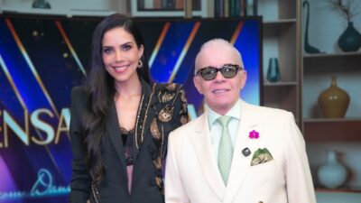 Daniela Albuquerque sorri ao lado de Chiquinho Scarpa de terno branco e óculos escuros no cenário do Sensacional