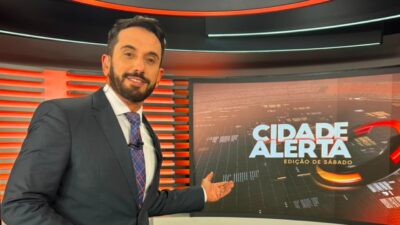 Dionisio Freitas de terno e gravata aponta para tela do Cidade Alerta - Edição de Sábado em estúdio da Record