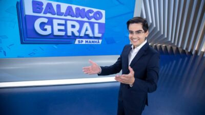 Dudu Camargo de terno sorri no estúdio do Balanço Geral SP Manhã com as mãos abertas diante do telão do programa