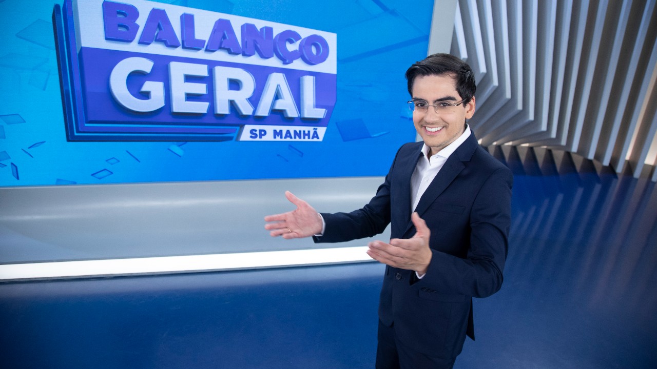 Dudu Camargo de terno sorri no estúdio do Balanço Geral SP Manhã com as mãos abertas diante do telão do programa