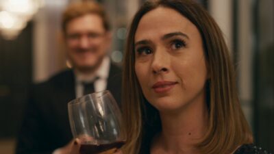 Brigitte (Tatá Werneck) segura taça de vinho enquanto Edvaldo (Guilherme Piva) aparece ao fundo em cena de Quem Ama Cuida