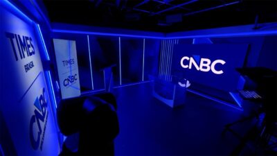 Estúdio da Times Brasil | CNBC com bancada de apresentação, telões exibindo logotipos, câmera em primeiro plano e iluminação azul ambiente