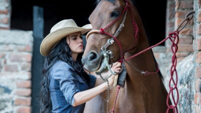 Fernanda Linhares (Livia Brito) de chapéu de cowboy segura as rédeas de um cavalo em estábulo de A Desalmada