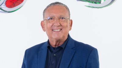 Galvão Bueno sorri de blazer azul e óculos diante de parede branca com bolas de futebol decorativas ao fundo
